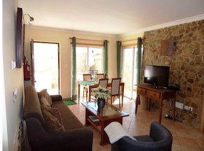 Un salon comprenant un canapé, une table basse avec un vase, un téléviseur posé sur un meuble en bois, et un coin repas avec une table et des chaises.