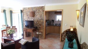 A living room with a sofa, a TV on a wooden stand, a dining area with a table and chairs, and a kitchen visible through an open doorway. - Casa Goncalito Ground floor (Bordeira)