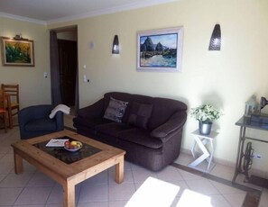 A living room with a dark brown sofa, a wooden coffee table with fruits, a painting on the wall, and a potted plant.