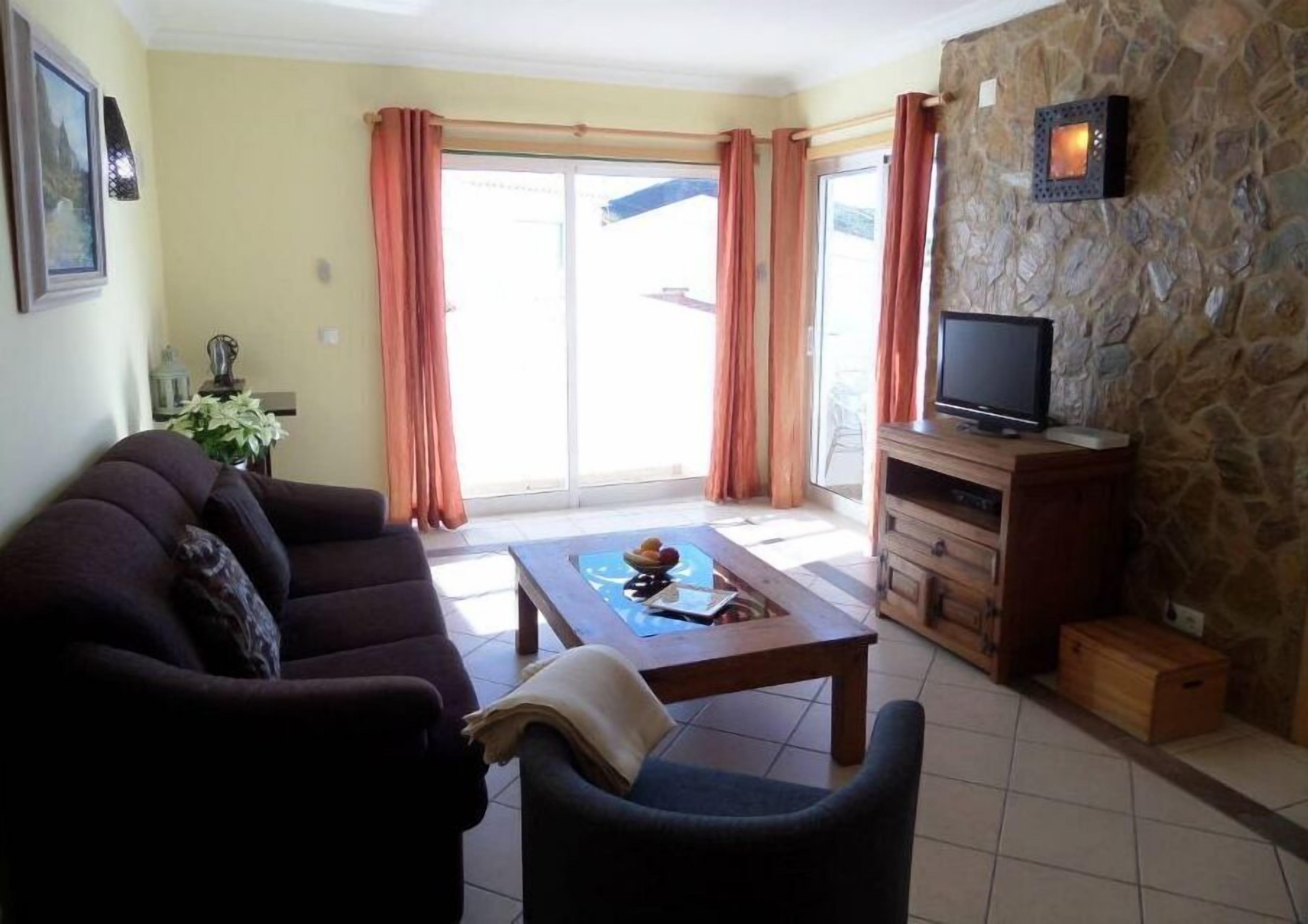 A living room with a sofa, a coffee table, a TV on a wooden cabinet, and a stone wall.