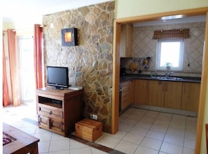 A kitchen with stone walls, wooden cabinets, a tiled floor, a television on a wooden stand, and a window with curtains.