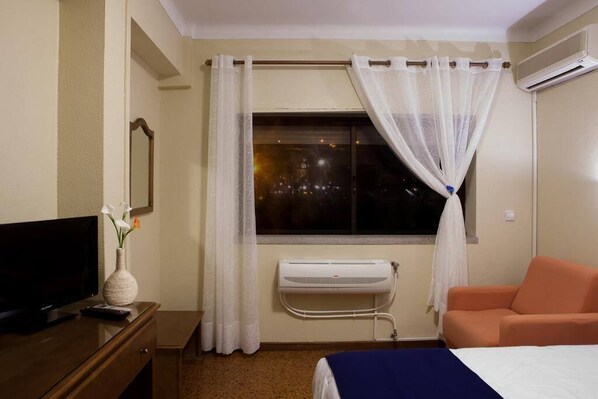 A hotel room with a bed, a TV, a bedside table with a vase, a window with curtains, and an air conditioning unit. - A Muralha (Almeida)