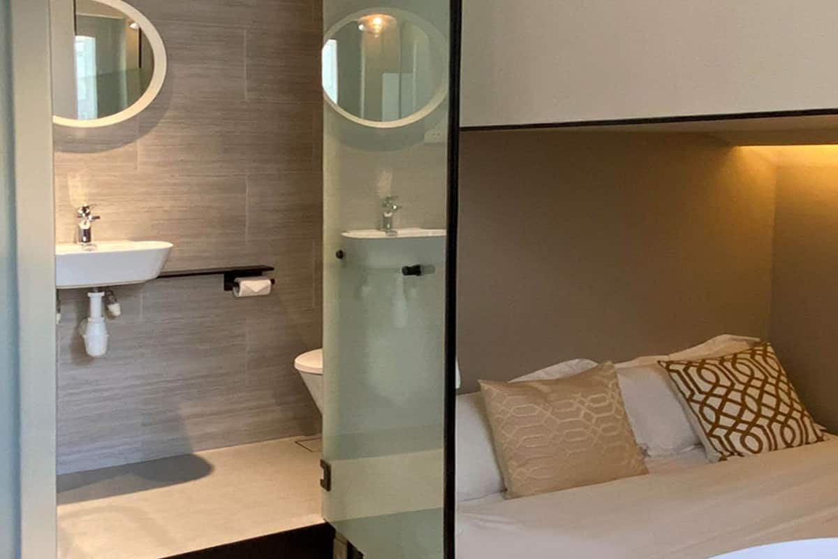 A modern bathroom with a sink, mirror, and toilet. The room features a bed with patterned pillows and a bedside table.