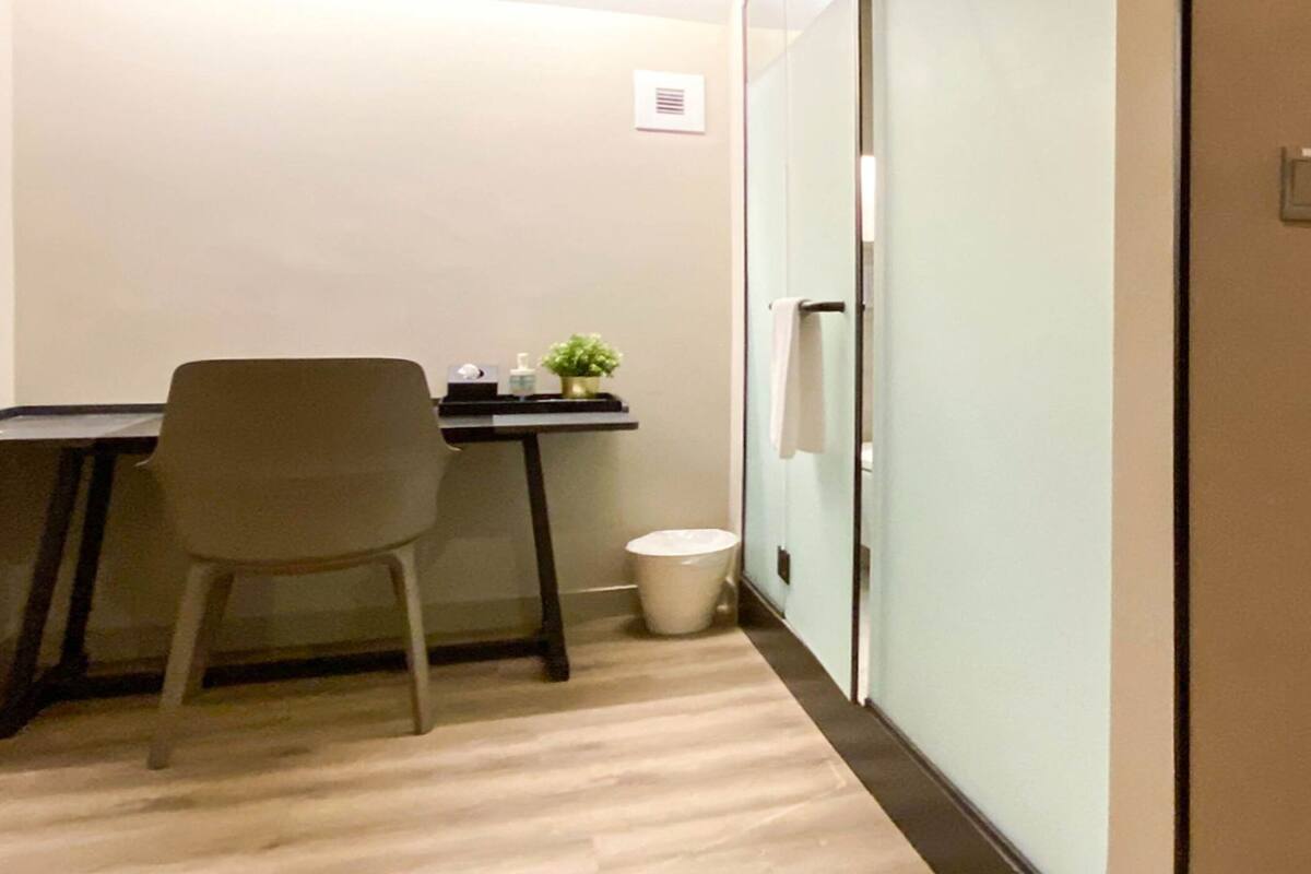 A hotel room with a bed, a desk, a chair, a small plant, and a trash bin.