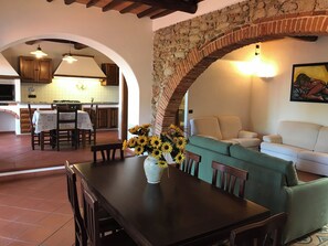 Un comedor con una mesa, sillas y un jarrón de girasoles. Se observa una cocina al fondo con una mesa y sillas.