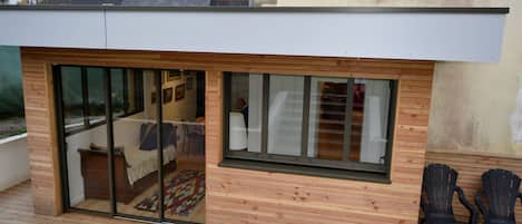 Ein modernes Gebäude mit Holzfassade, große Schiebeglastüren, die zu einem Raum mit einem Bett und einem Teppich führen, und zwei schwarze Stühle davor.