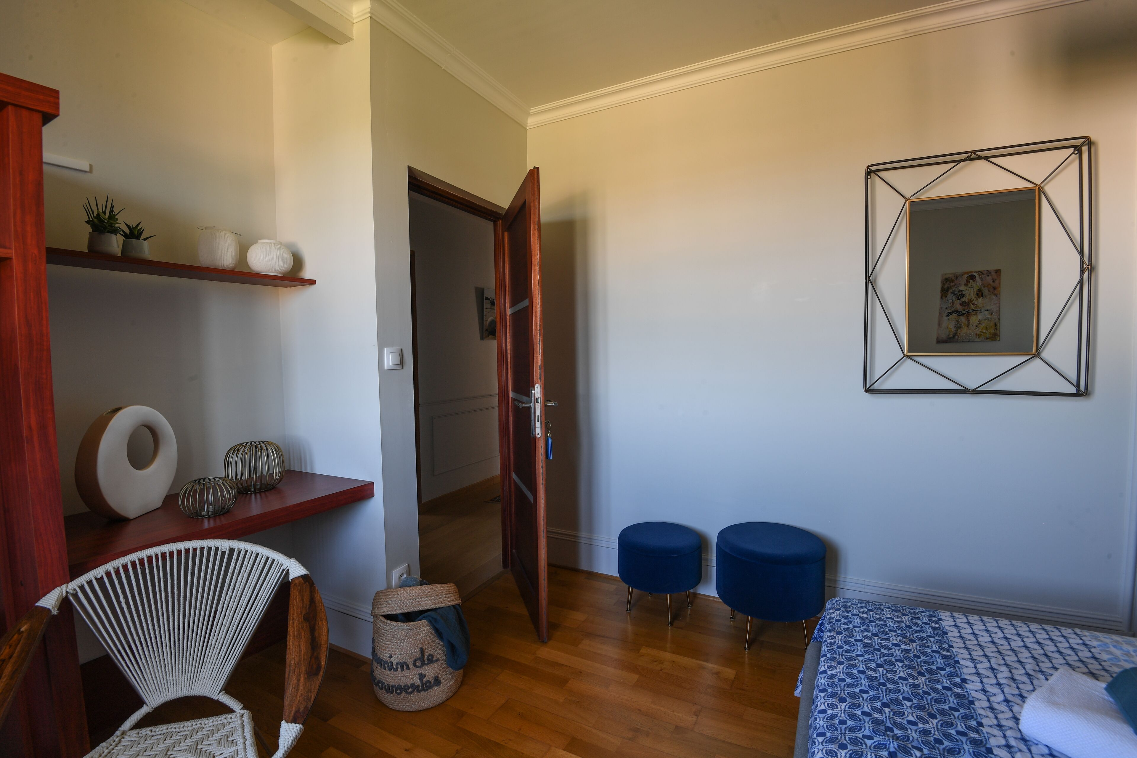 Une pièce comprenant un lit, deux poufs bleus, un fauteuil en osier, une étagère en bois ornée d’objets décoratifs et une porte donnant sur une autre pièce.