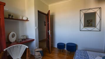 Ein Zimmer mit einem Bett, zwei blauen Poufs, einem Korbstuhl, einem Holzregal mit Dekoration und einer Tür zu einem weiteren Raum.