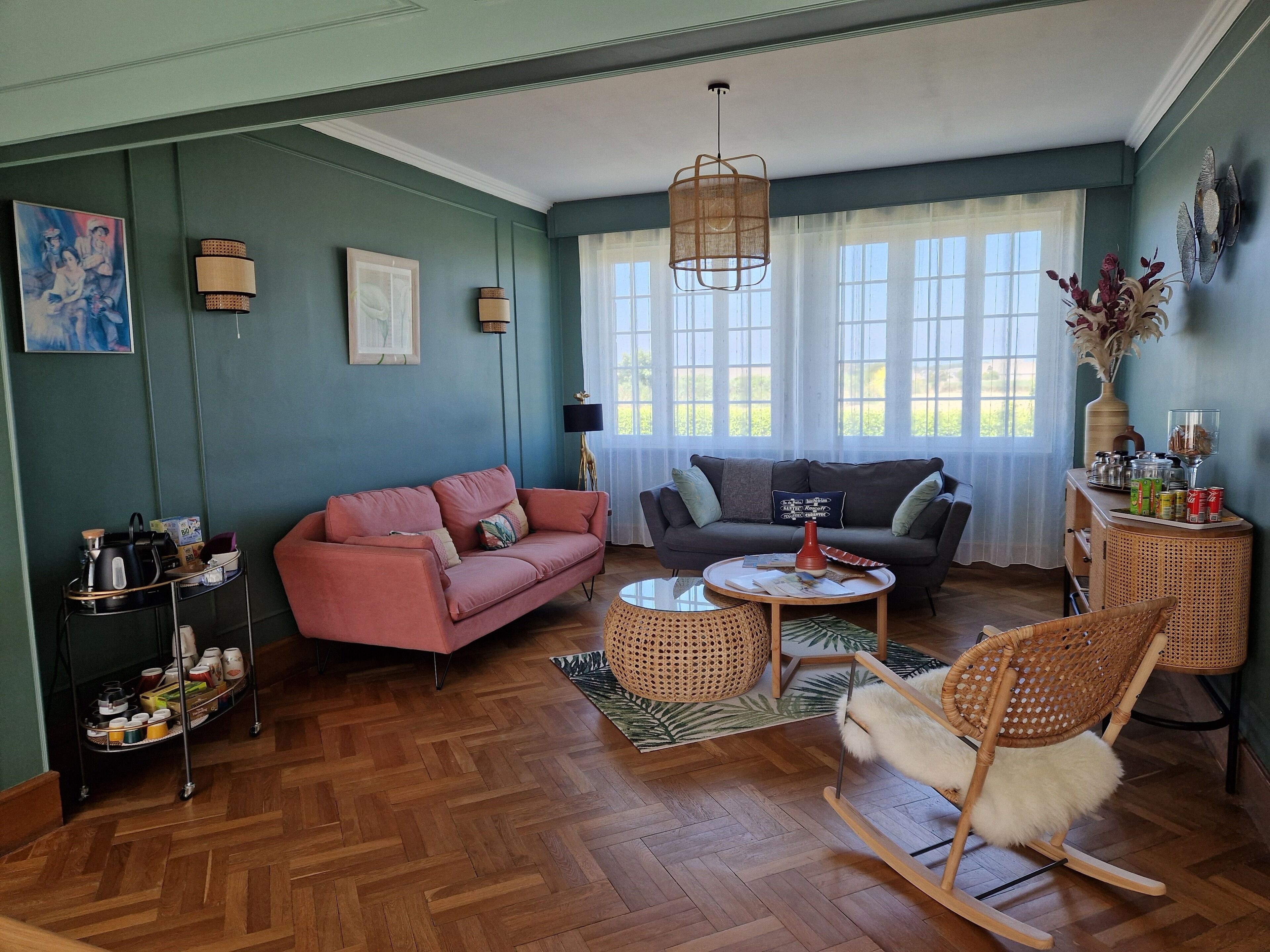 A cozy living room with a herringbone wood floor, a pink sofa, a grey sofa, a round coffee table, and a wicker rocking chair.