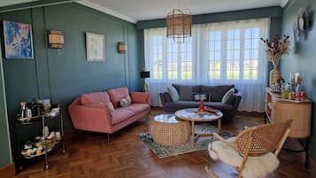 Een gezellige woonkamer met een visgraat houten vloer, een roze bank, een grijze bank, een ronde salontafel en een rieten schommelstoel.