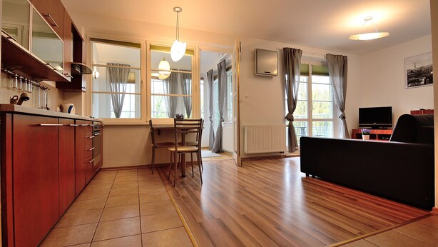 A modern kitchen with wooden cabinets, a dining area with a table and chairs, a black sofa, and a TV in a living room. - Victus Apartamenty - Petit (Sopot)