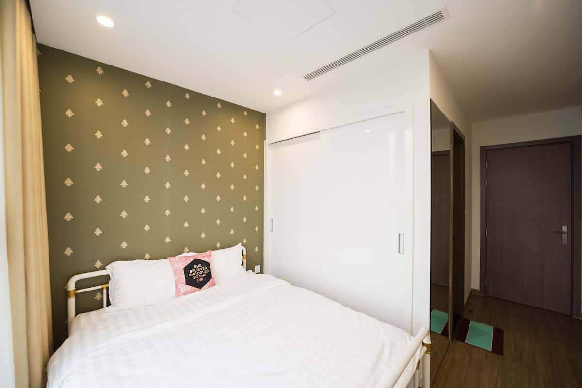 A neatly made bed with a white comforter and pillows, a green patterned wall, a white door, and a wooden floor.