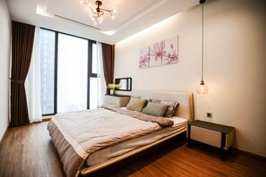 A modern bedroom with a large bed, a bedside table, a nightstand, a wall-mounted light, and a window with curtains. - Homefromhome Skylake Apartment (Hanoi)