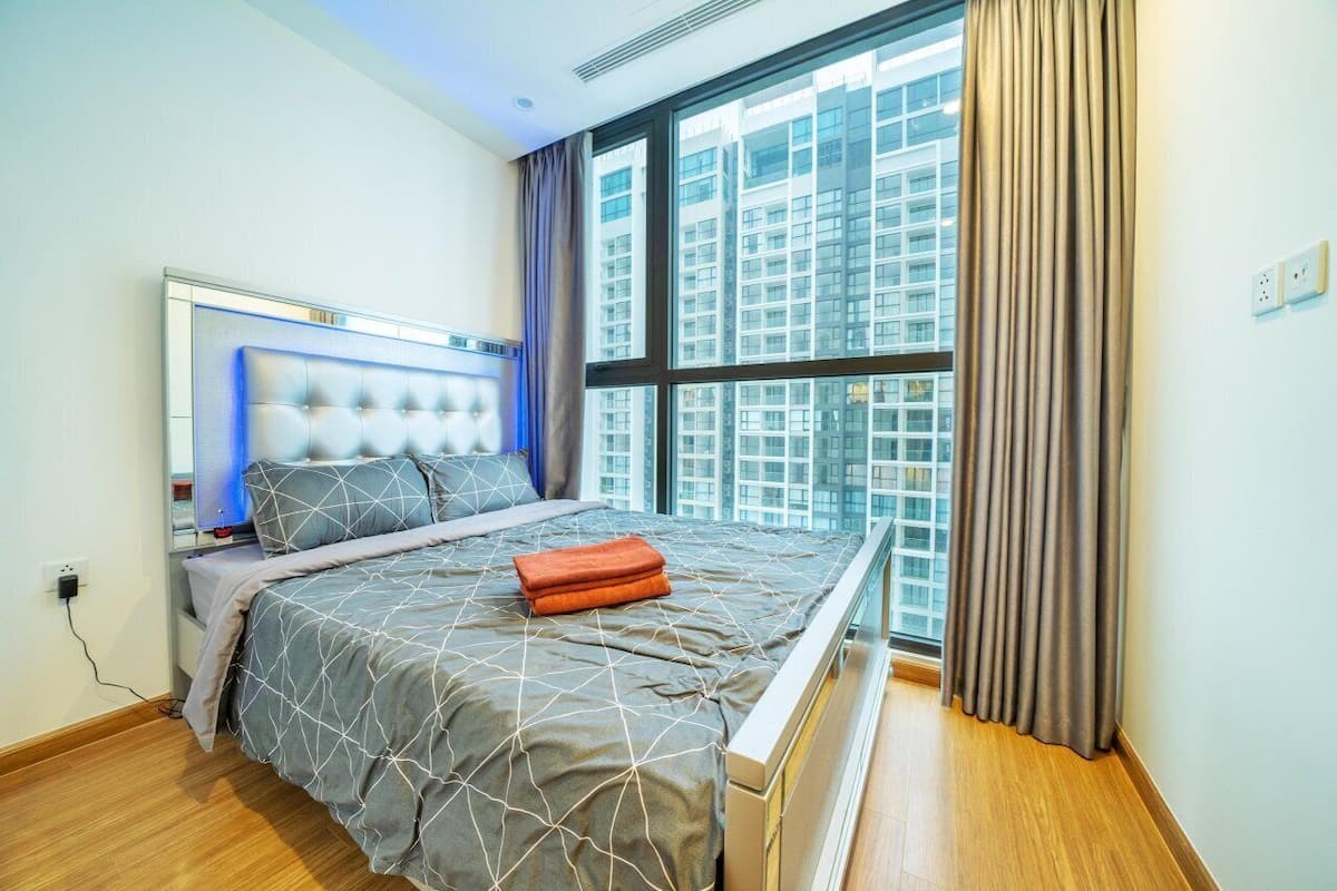 A modern bedroom with a large bed, a headboard with built-in lighting, a window with curtains, and a view of high-rise buildings.