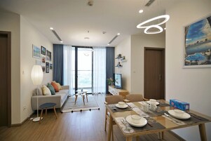 Una sala de estar moderna con sofá, mesa de comedor y televisor. Se aprecia un ventanal con cortinas, una lámpara y un cuadro en la pared.