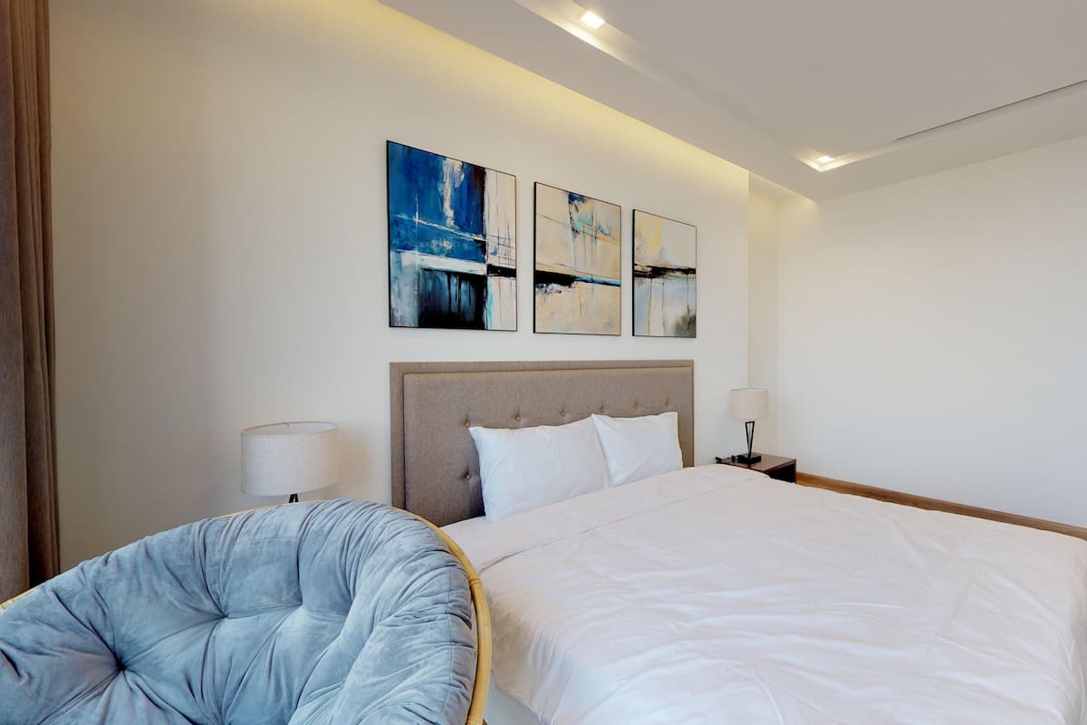 A modern hotel room with a large bed, a tufted armchair, and three abstract paintings on the wall.