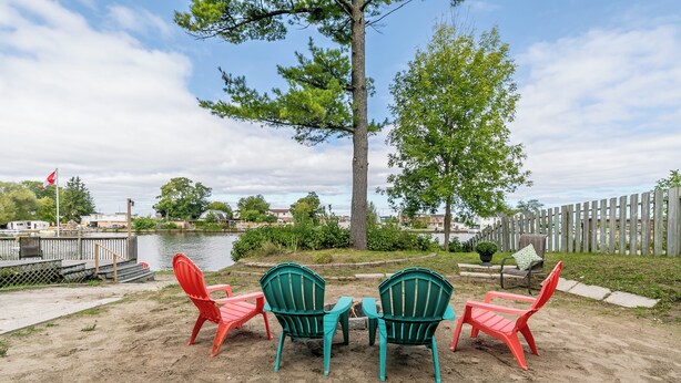 A sandy outdoor seating area with two green and two red Adirondack chairs, a small table, and a flagpole. - Beach1 Riverfront Resort - #32 - Wasaga Beach (Wasaga Beach)