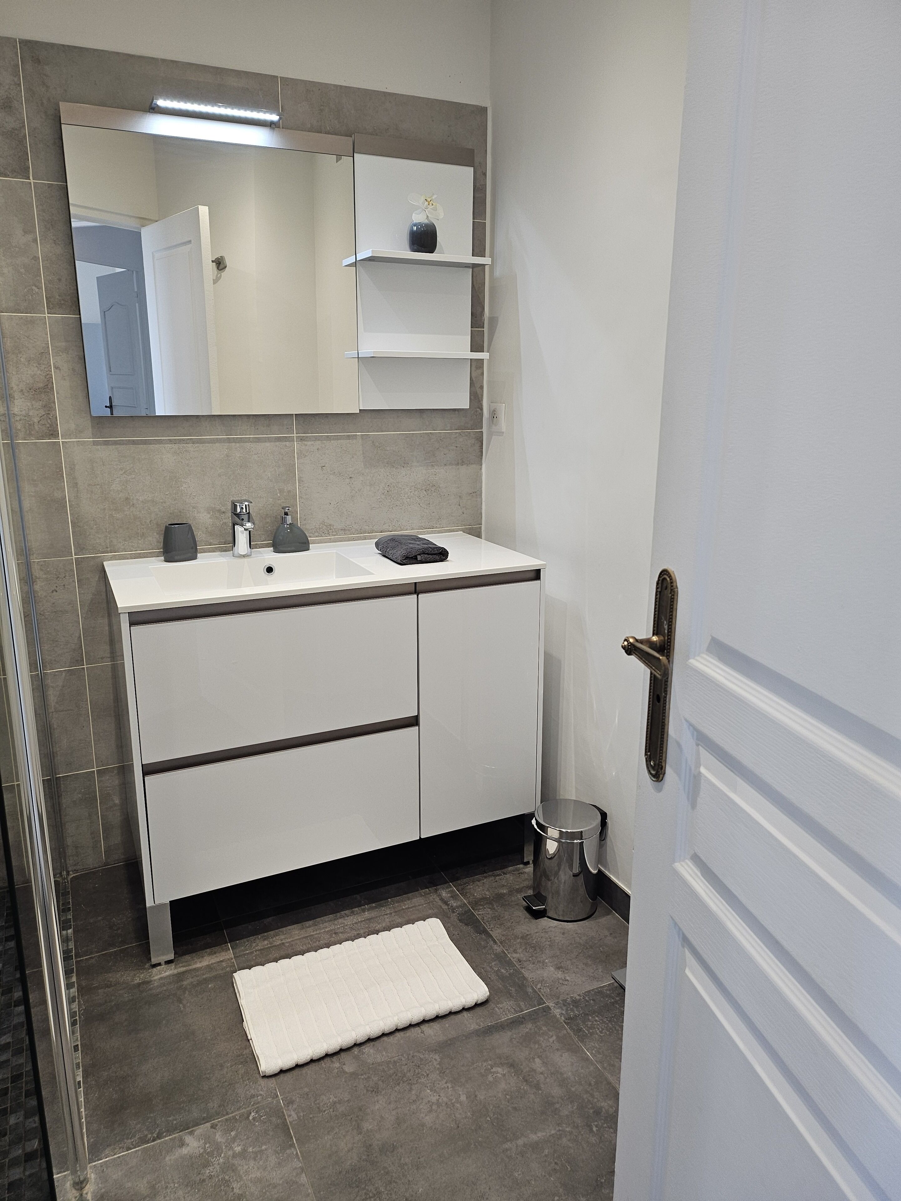 Un baño con tocador blanco, un espejo, un estante pequeño con un florero y un tapete de baño blanco en el suelo.