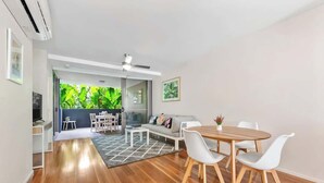 A modern living room with a wooden floor, white furniture, a dining table, and a view of a balcony with greenery.