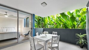 A dining area with a white table and chairs, a vase with flowers, and a view of lush green plants through a glass partition.