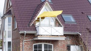 A two-story house with a red-tiled roof, a dormer window, and a small balcony with a yellow awning. - Seetiger (Krummhoern)
