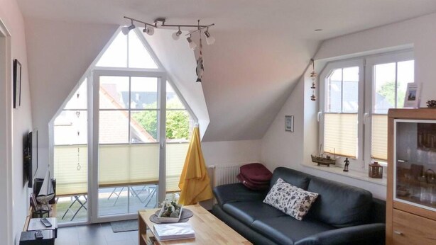 A modern living room with a black leather sofa, a wooden coffee table, and a TV mounted on the wall. There is a balcony with a yellow towel and a red cushion. - Seetiger (Krummhoern)