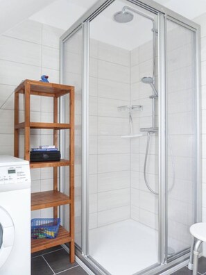 A shower with a glass door, a wooden shelf with items, and a washing machine. - Seetiger (Krummhoern)