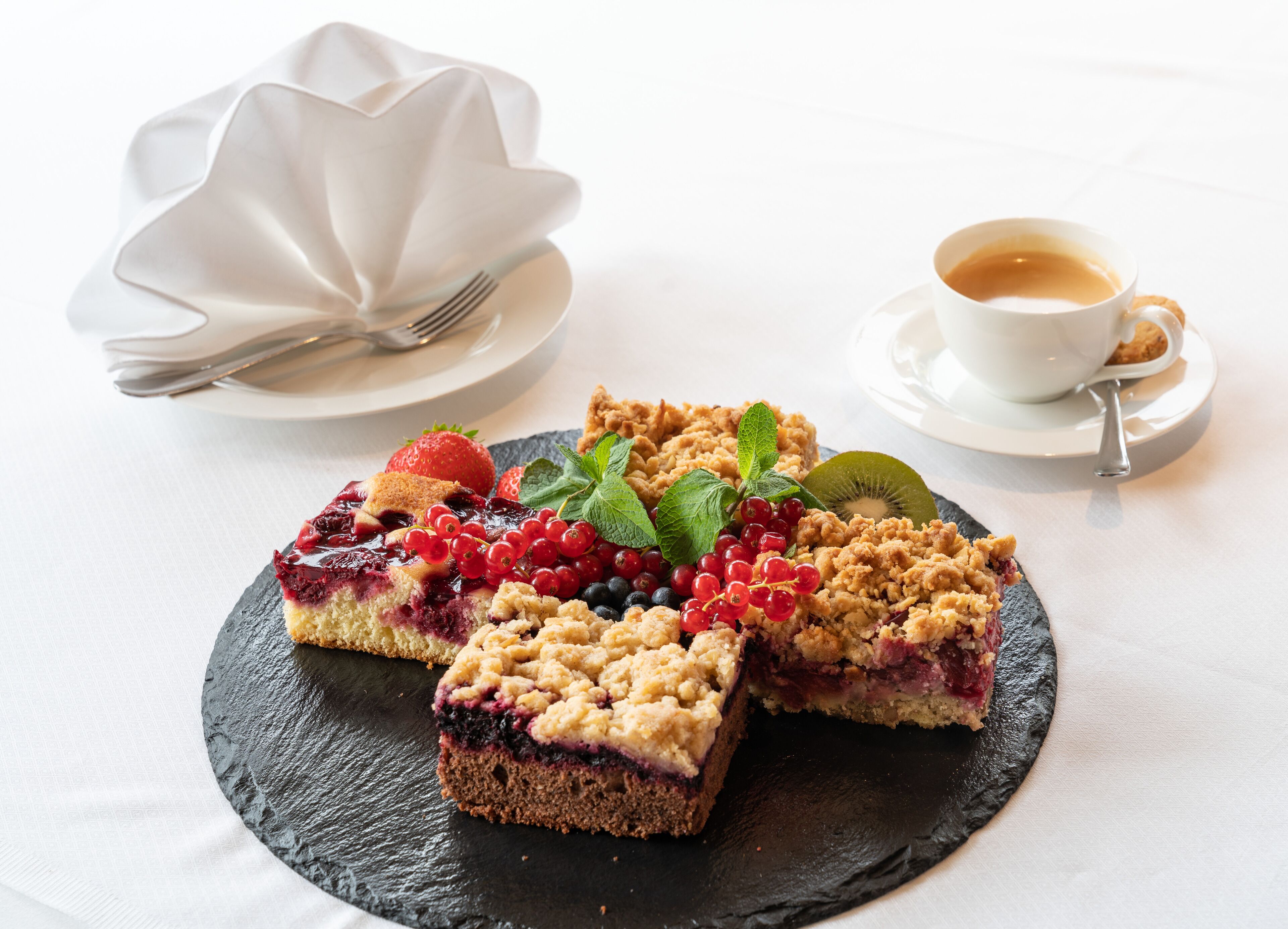 Une assiette à dessert avec une part de gâteau garnie de fruits rouges et de miettes, une tasse de café et une serviette blanche avec une fourchette, le tout posé sur une nappe blanche.