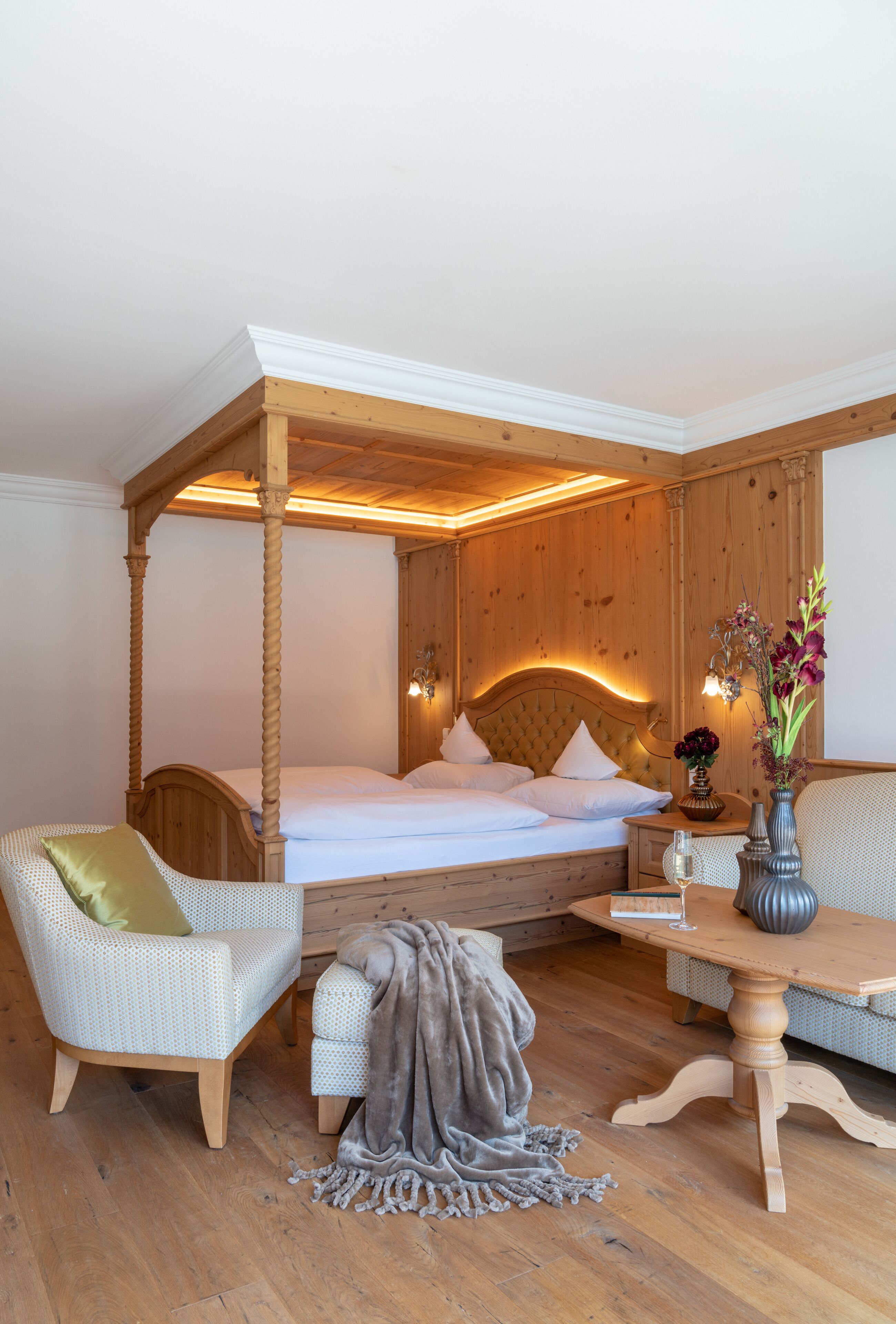 A four-poster bed with a wooden headboard, a bedside table with a vase, and a seating area with a sofa and armchair.