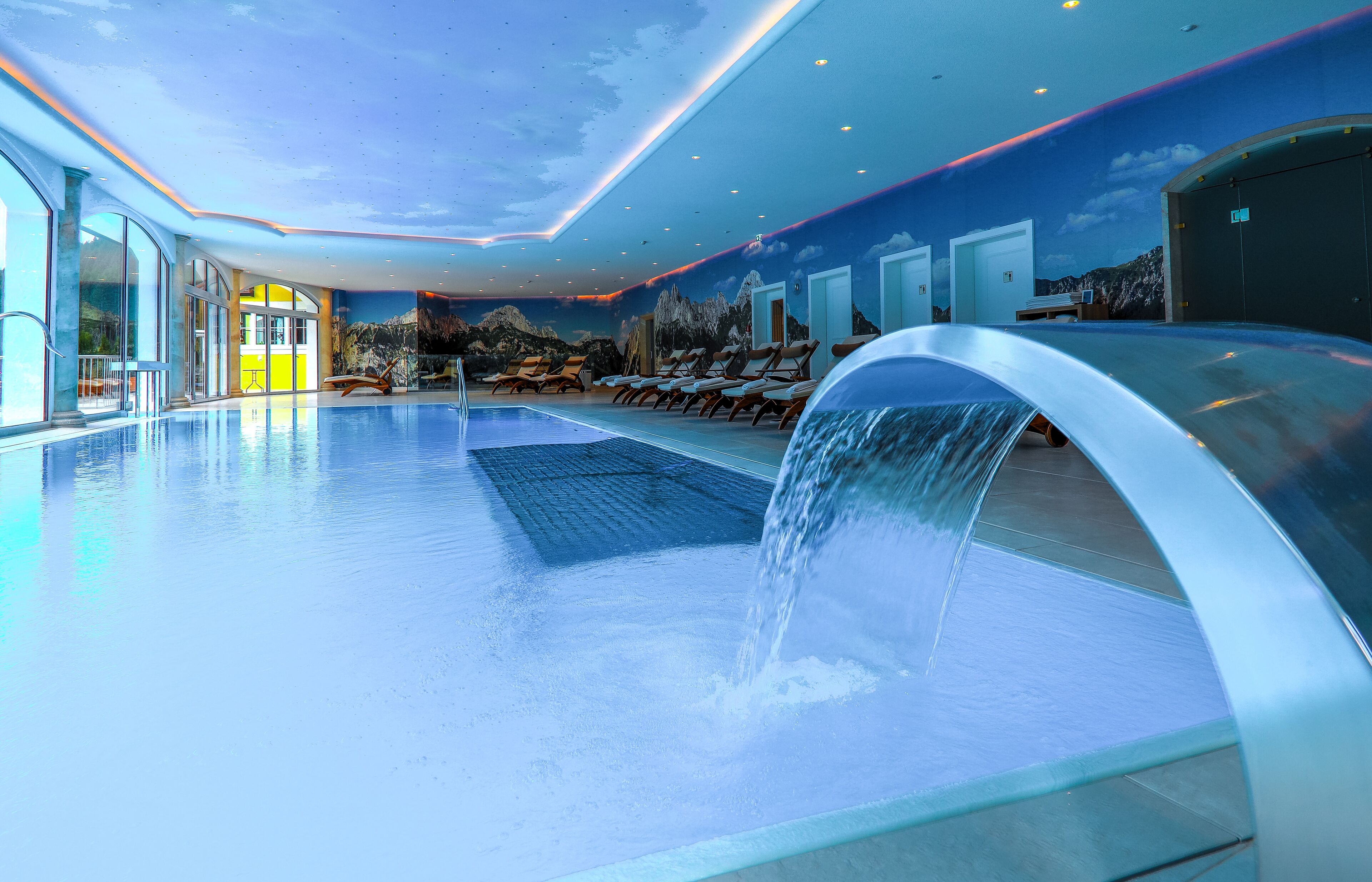 Ein moderner, überdachter Pool mit Wasserfall, Liegestühlen und einer Wandmalerei einer Berglandschaft.