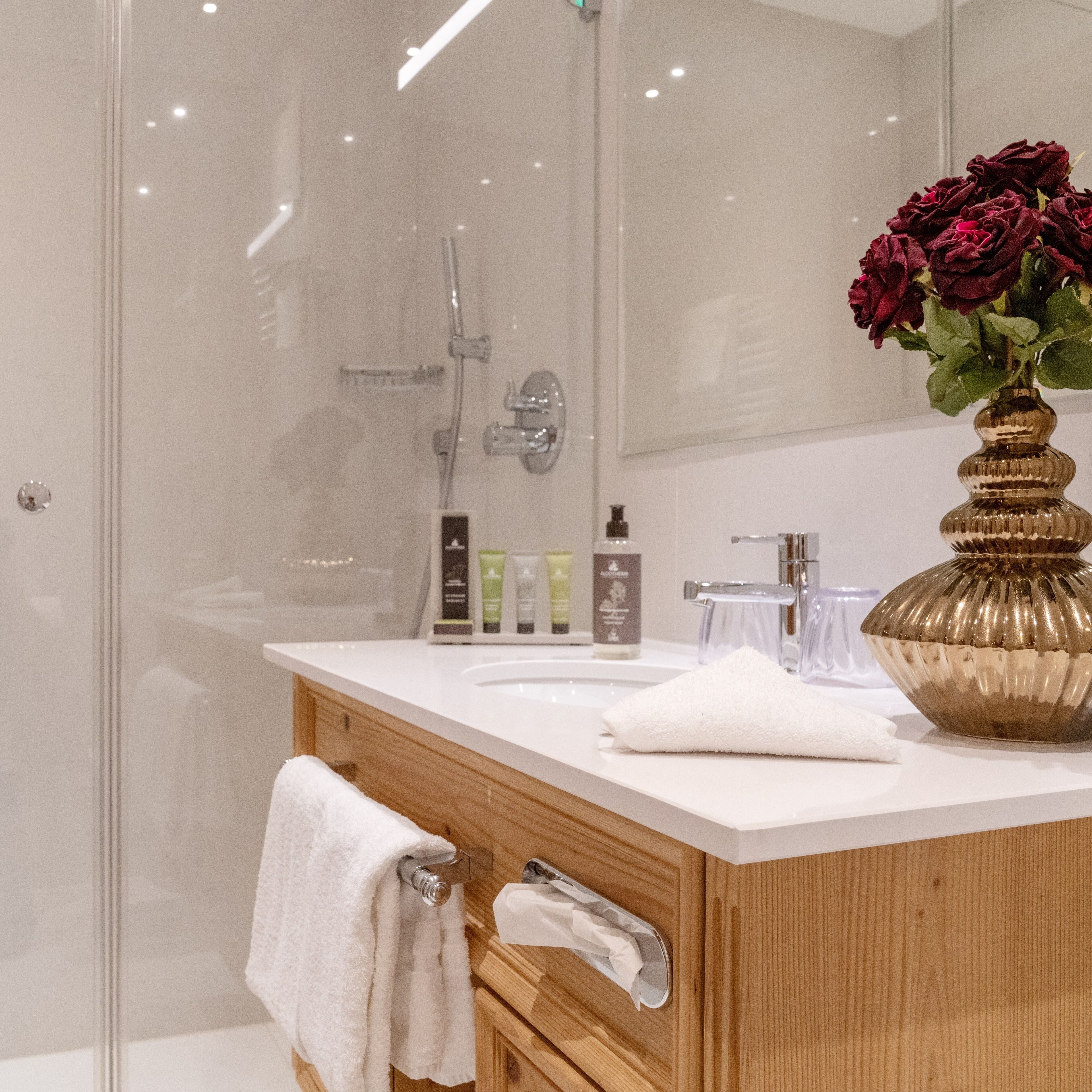Un baño moderno con mampara de vidrio, tocador de madera con un jarrón dorado y una toalla blanca colgada en la puerta.