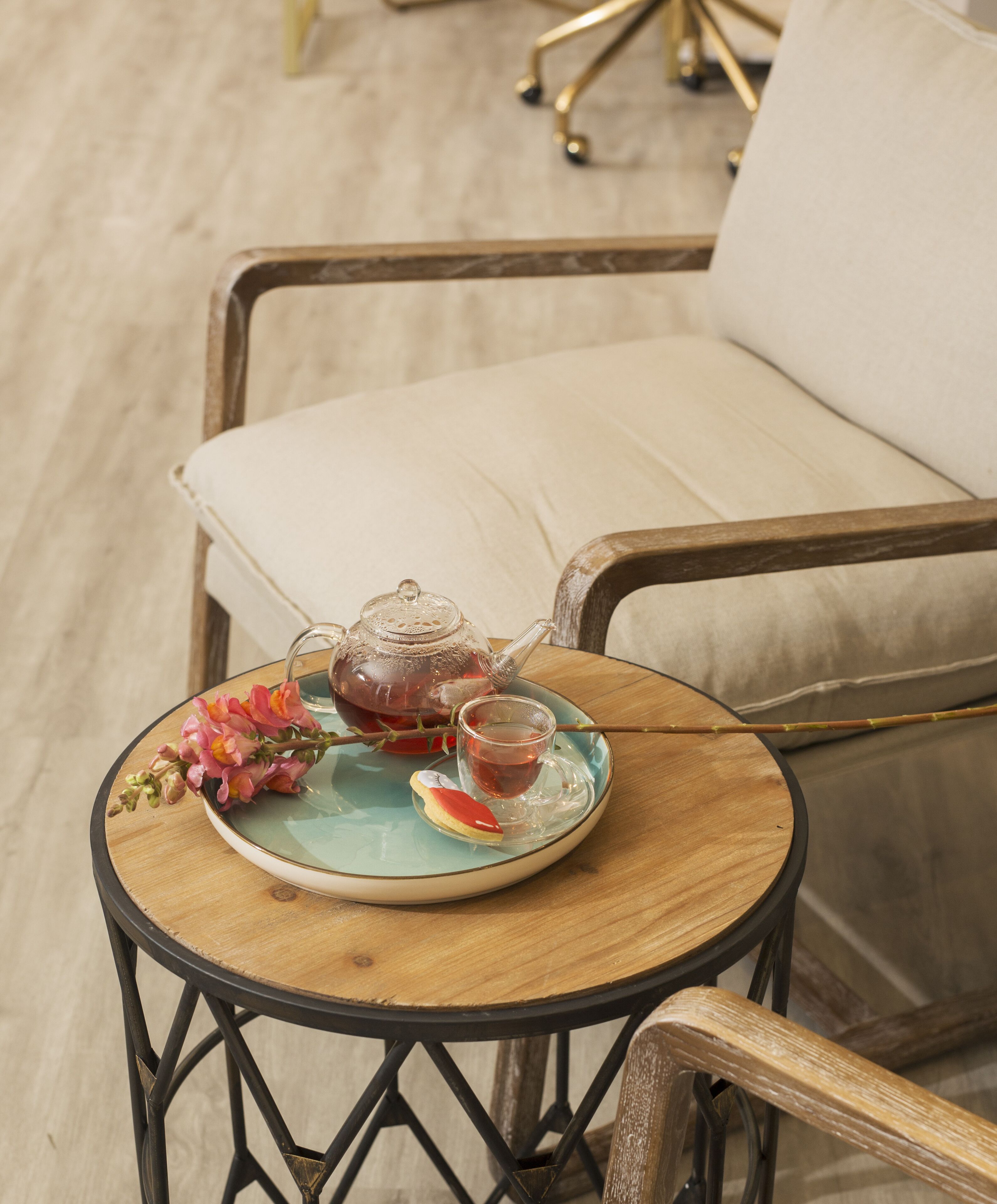 Une table ronde en bois, sur laquelle se trouvent une théière et une tasse en verre, une assiette avec une tranche de fruit et un bouquet de fleurs.