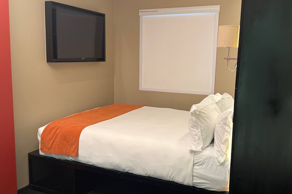 Un petit lit bien fait, avec une literie blanche et orange, une tête de lit noire et un téléviseur fixé au mur.