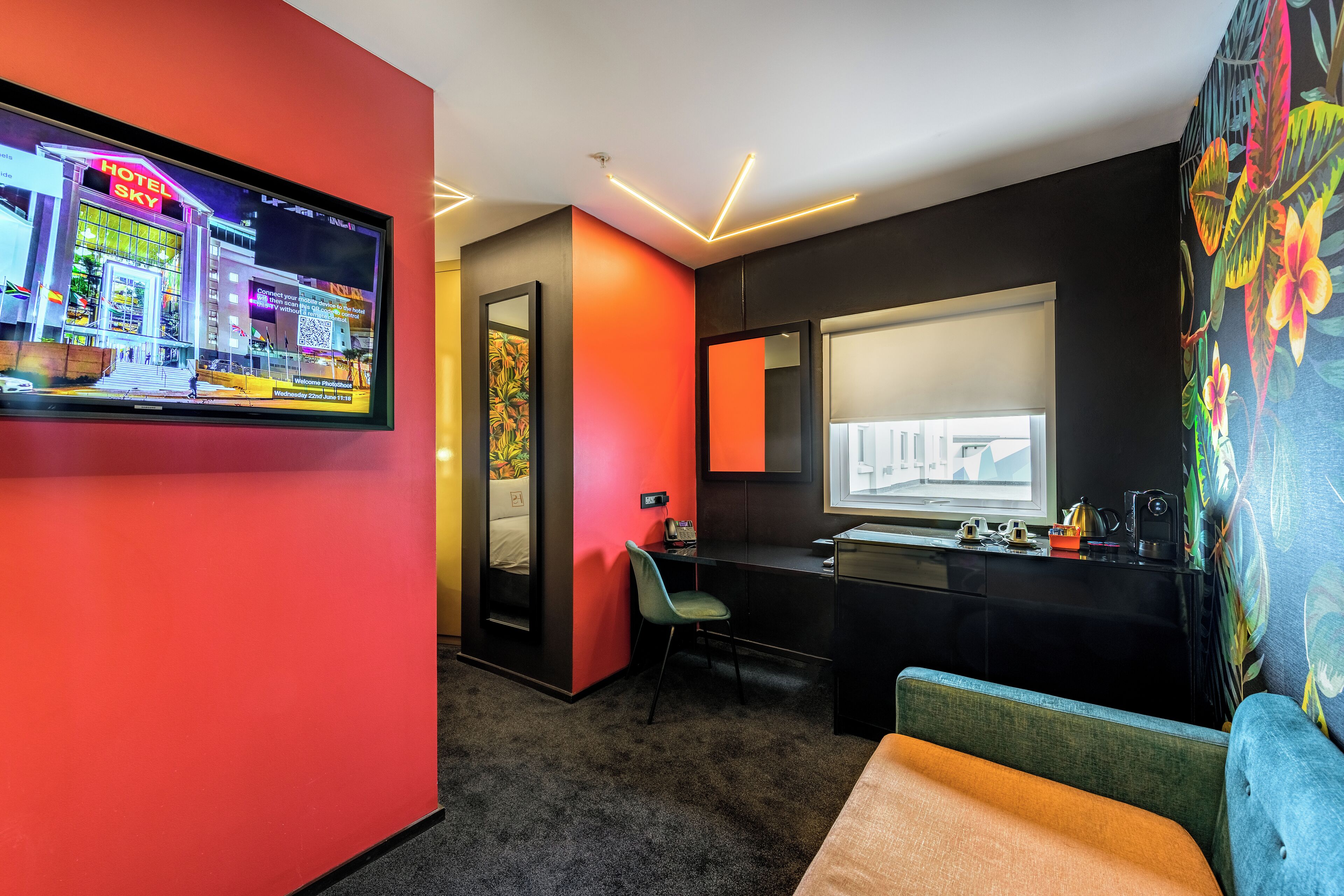 A modern hotel room with a TV displaying a hotel scene, a desk with a chair, a large window, and a colorful wall mural.