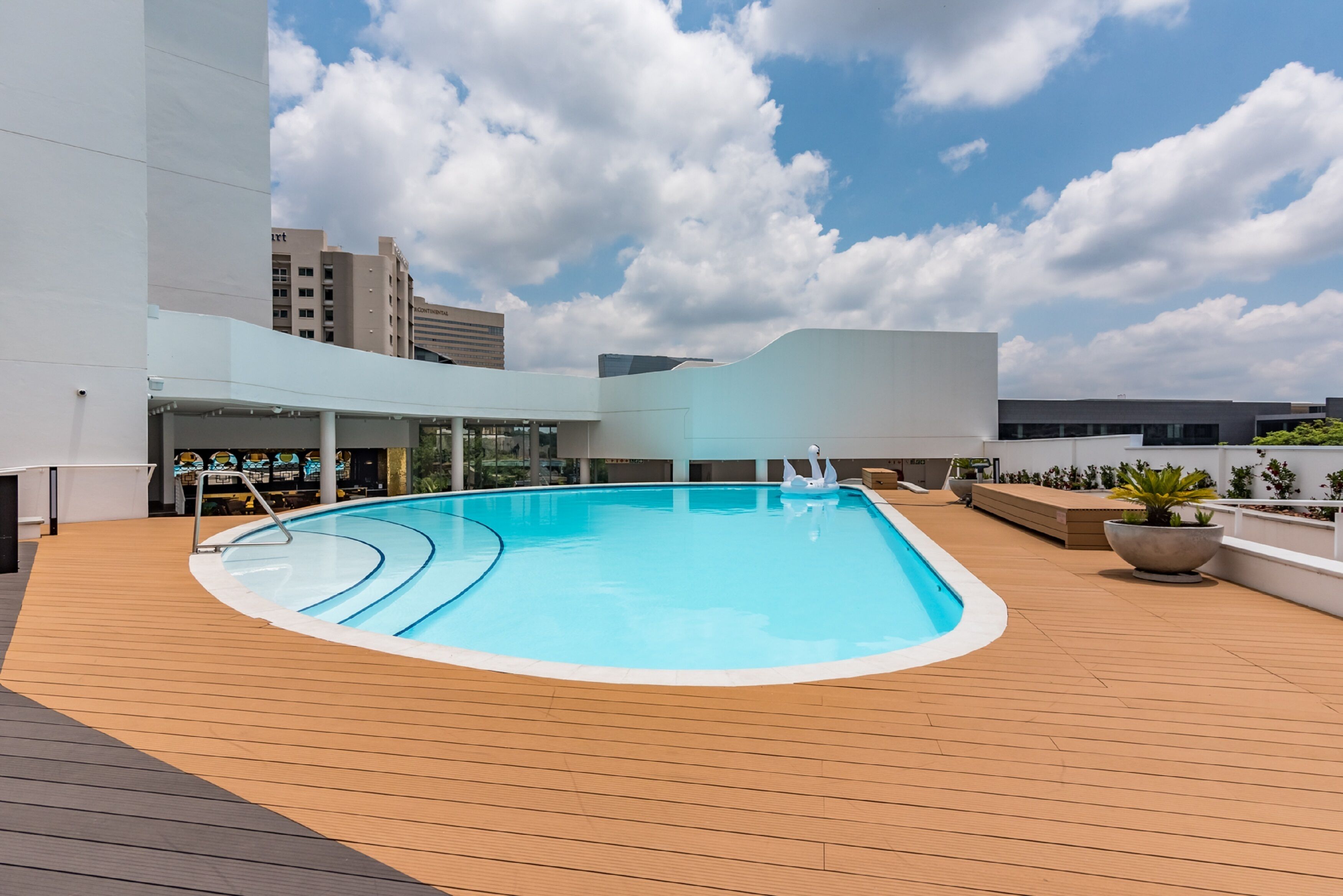Une piscine sur le toit, dotée d’une terrasse en bois, entourée de bâtiments modernes et surplombant un ciel dégagé.