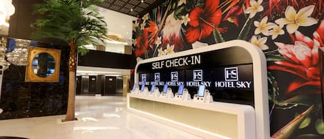 A self-check-in kiosk with multiple screens, a large floral mural, a palm tree, and a mirror on the wall.