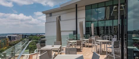 A rooftop terrace with outdoor seating, a glass facade building, and a city view.