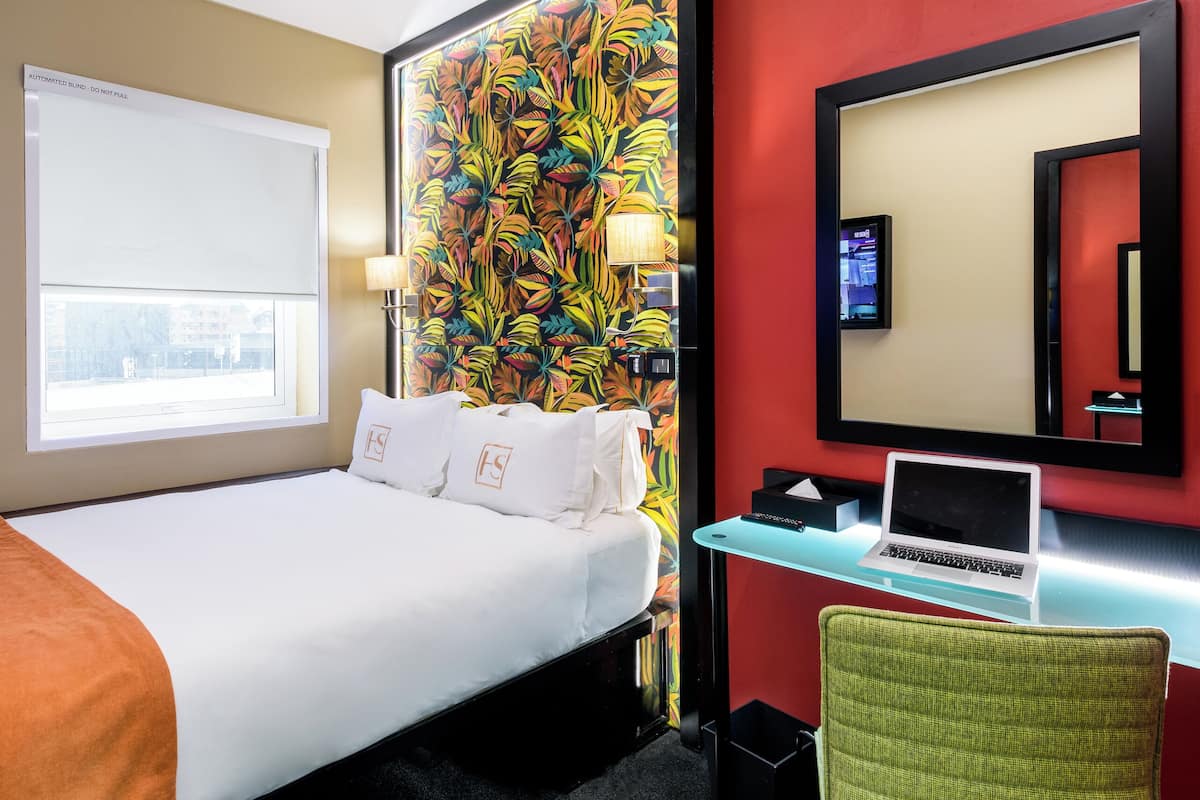 Une chambre d’hôtel avec un lit, un bureau avec un ordinateur portable, une chaise, un grand miroir et une décoration murale colorée.