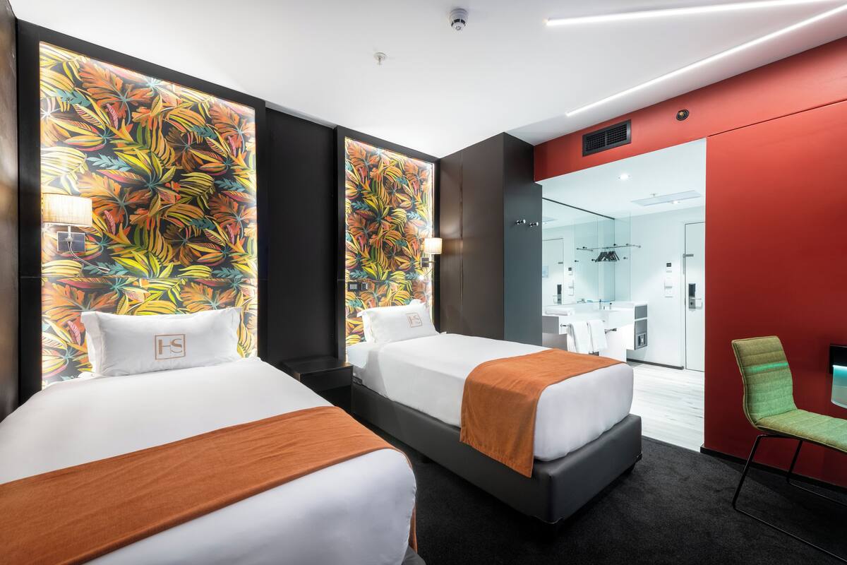 Une chambre d’hôtel avec deux lits, un mur d’accent rouge, une grande fenêtre et une salle de bain visible à travers la porte en verre.