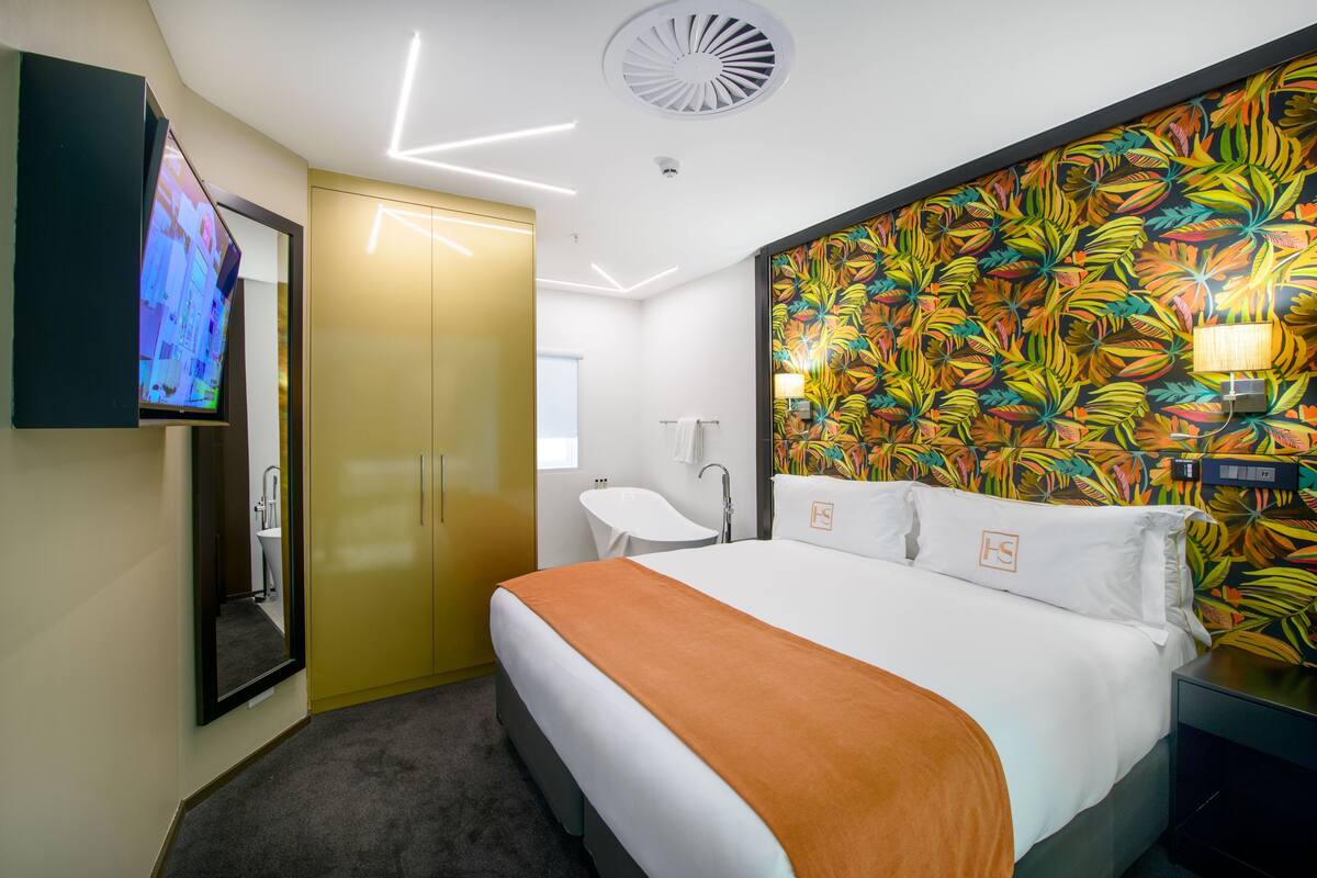 Une chambre d’hôtel moderne dotée d’un grand lit, d’une télévision, d’une salle de bain et d’un mur orné d’un motif de feuilles aux couleurs vives.
