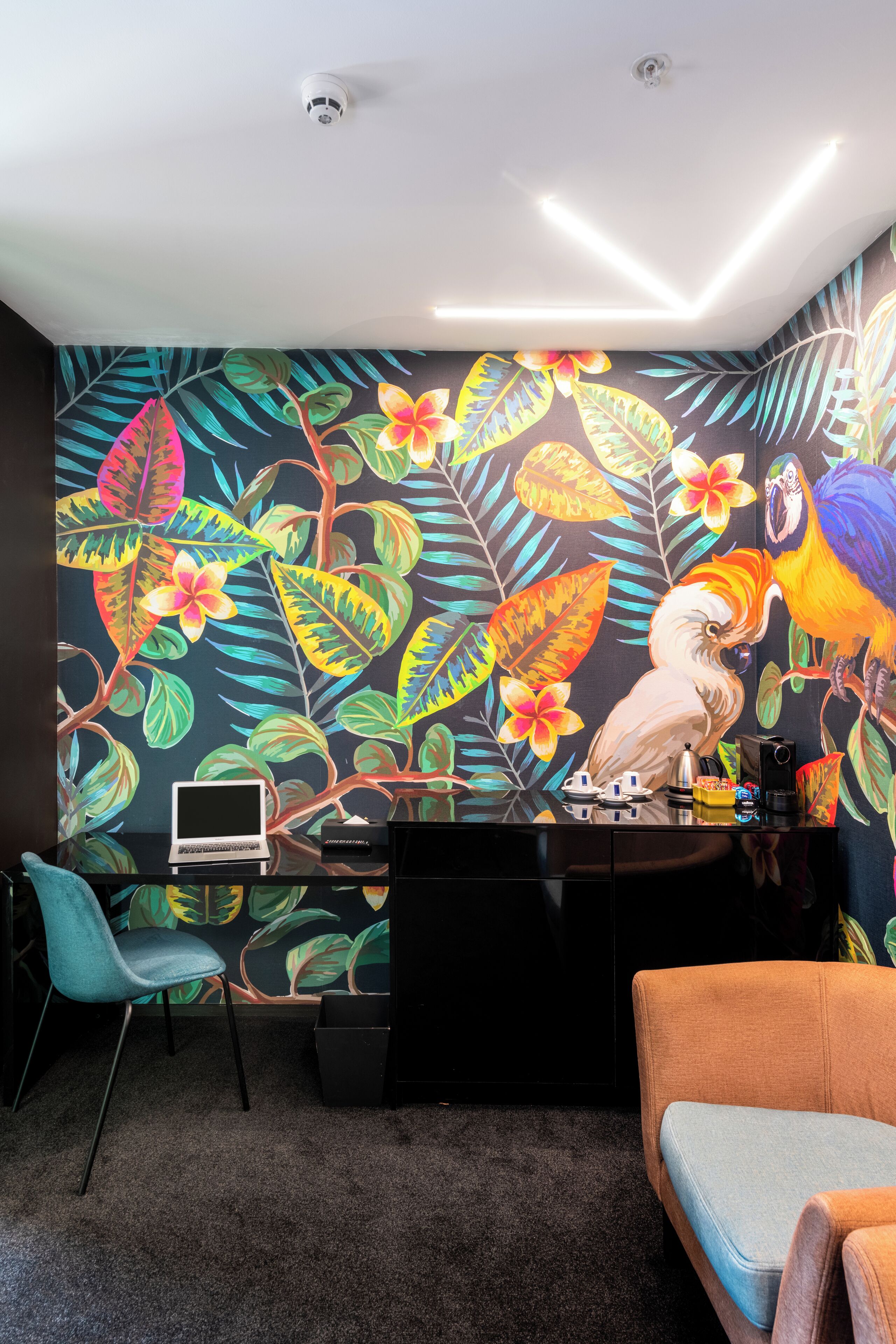 Una habitación con un vibrante mural tropical, un escritorio con una computadora portátil, una cafetera y una silla.