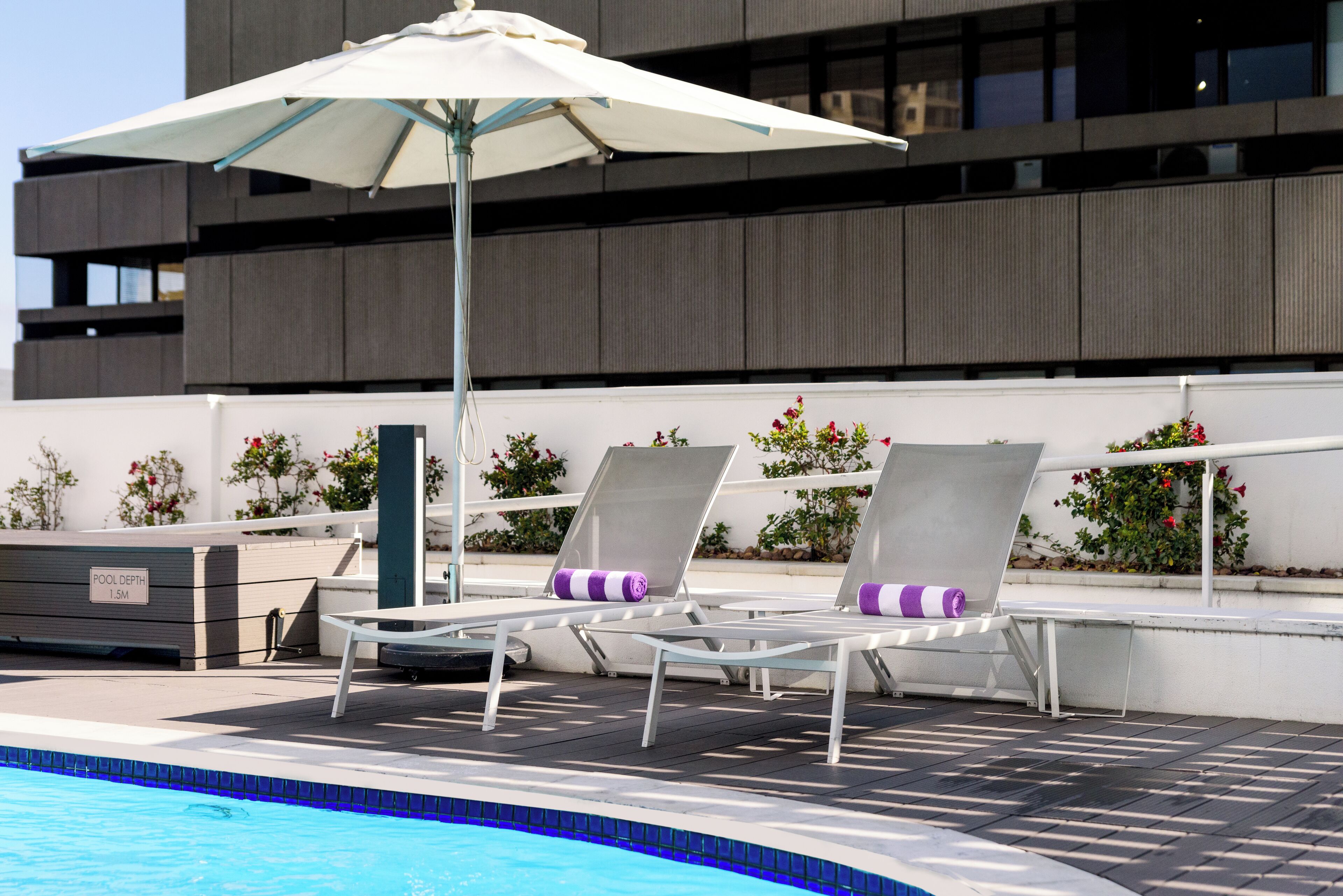 A pool area with lounge chairs and an umbrella, a sign indicating pool depth, and a modern building in the background.