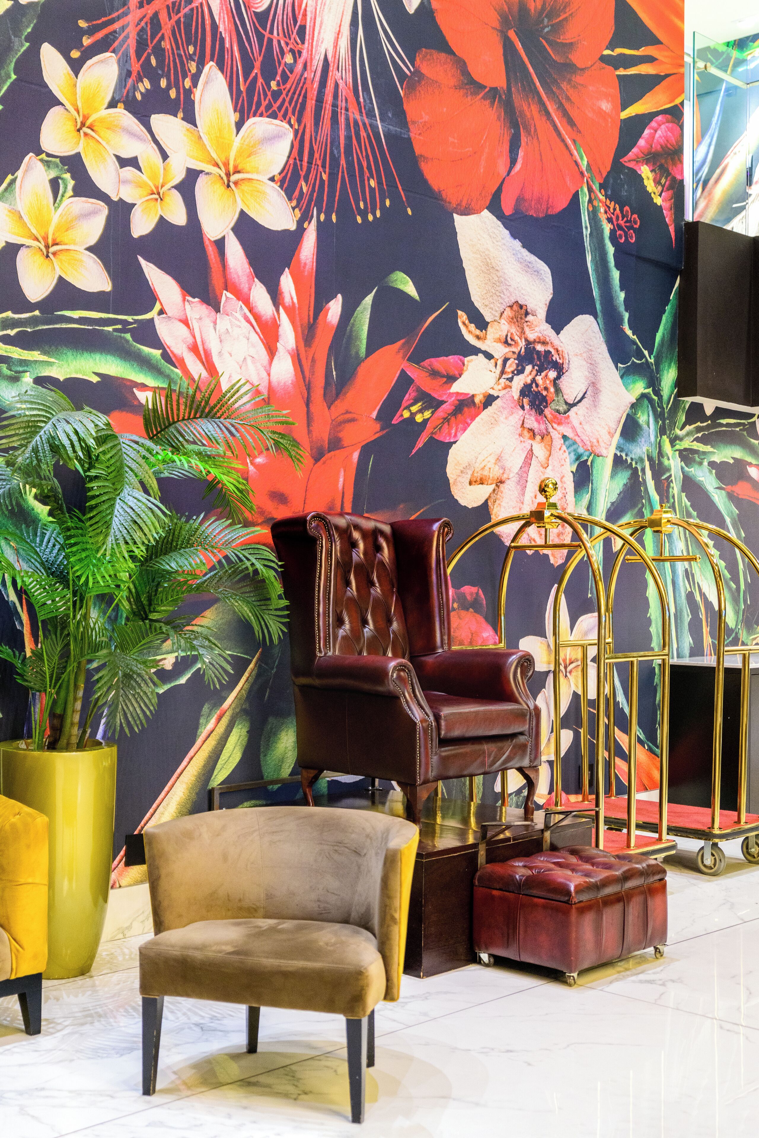 A room with a floral mural, a brown leather armchair, a matching ottoman, a gold-framed chair, and a potted plant.