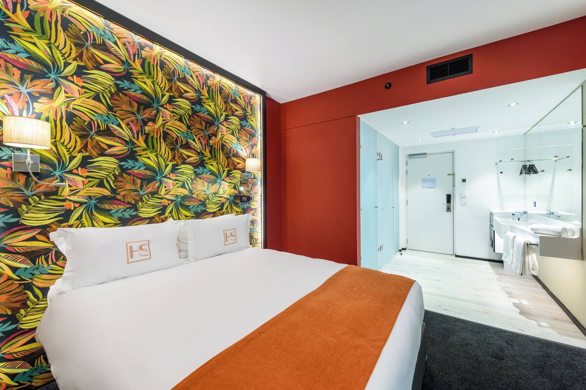 Une chambre d’hôtel avec un lit, un mur décoré d’un motif coloré représentant des feuilles, un mur d’accent rouge et une salle de bain visible à travers une porte ouverte.