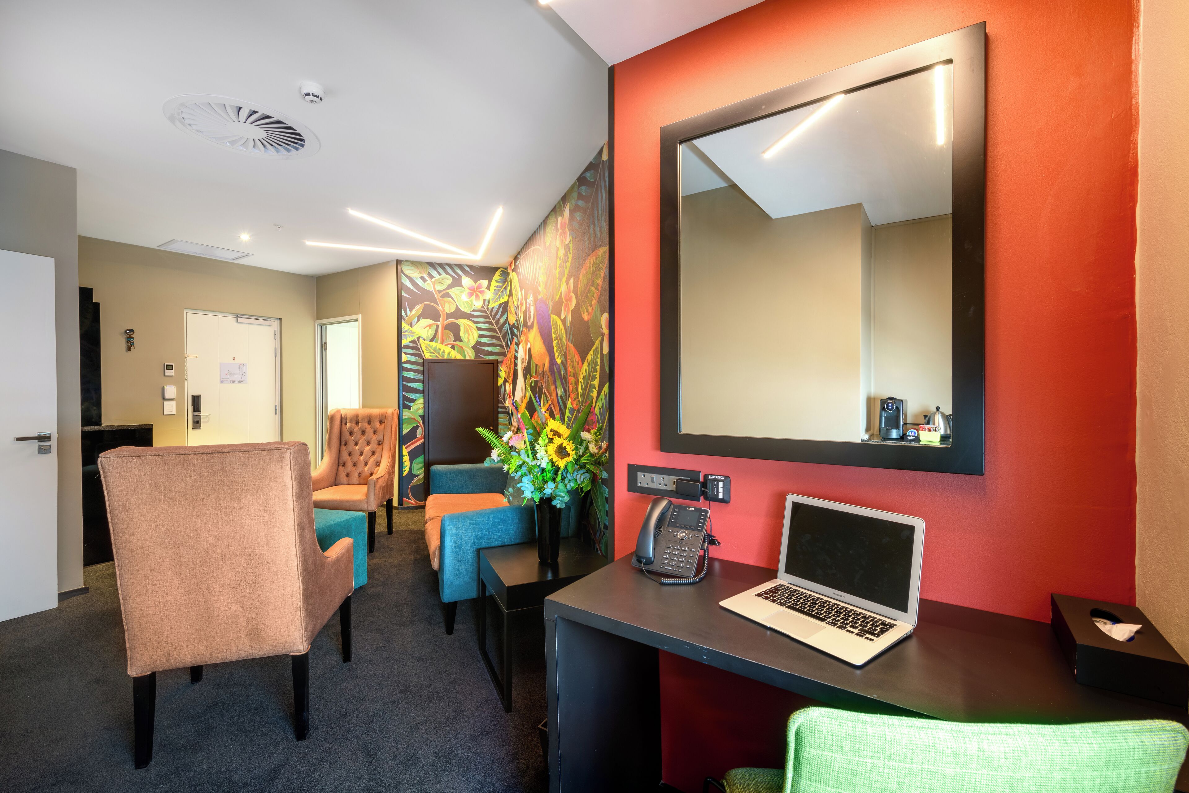 Une chambre d’hôtel équipée d’un bureau, d’un ordinateur portable, d’un téléphone et d’un miroir. On y trouve également des chaises et un vase avec des fleurs.