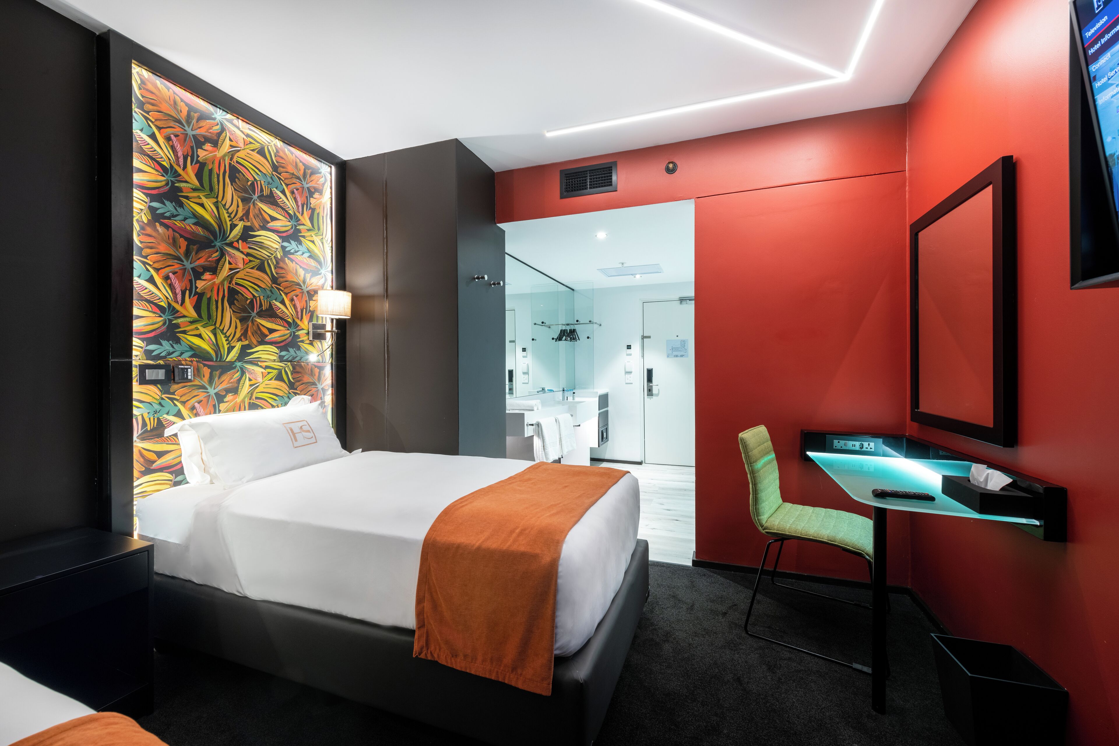 Una habitación de hotel moderna con una cama, un escritorio con una computadora, una silla y un baño visible a través de una puerta abierta.