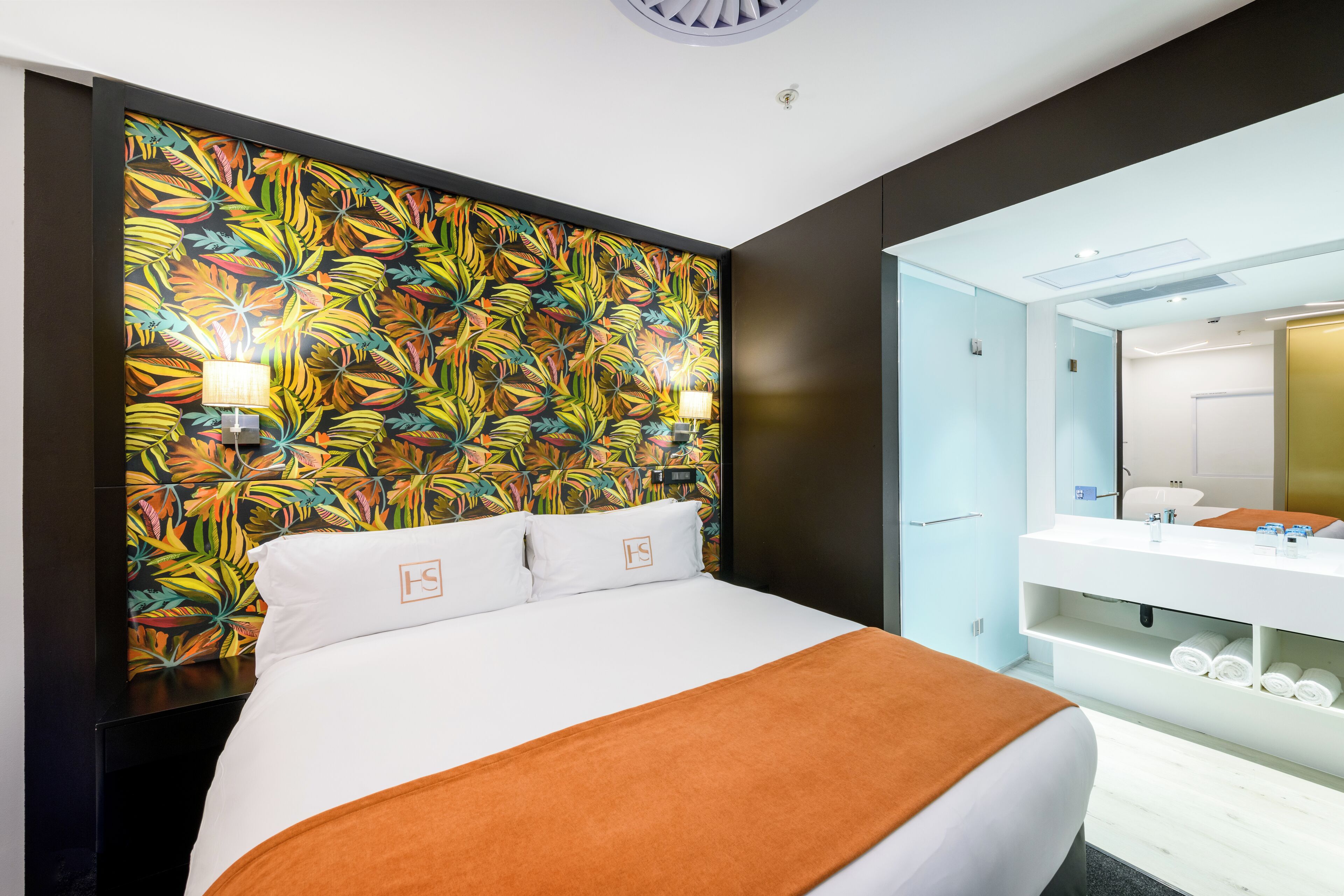 Une chambre d’hôtel avec un grand lit, un mur orné d’un motif de feuilles tropicales, une salle de bain avec un lavabo et un miroir blancs, et un luminaire au plafond.
