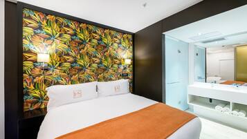 Una habitación de hotel con una cama grande, una pared con motivos de hojas tropicales, un baño con lavabo y espejo blancos, y una lámpara en el techo.