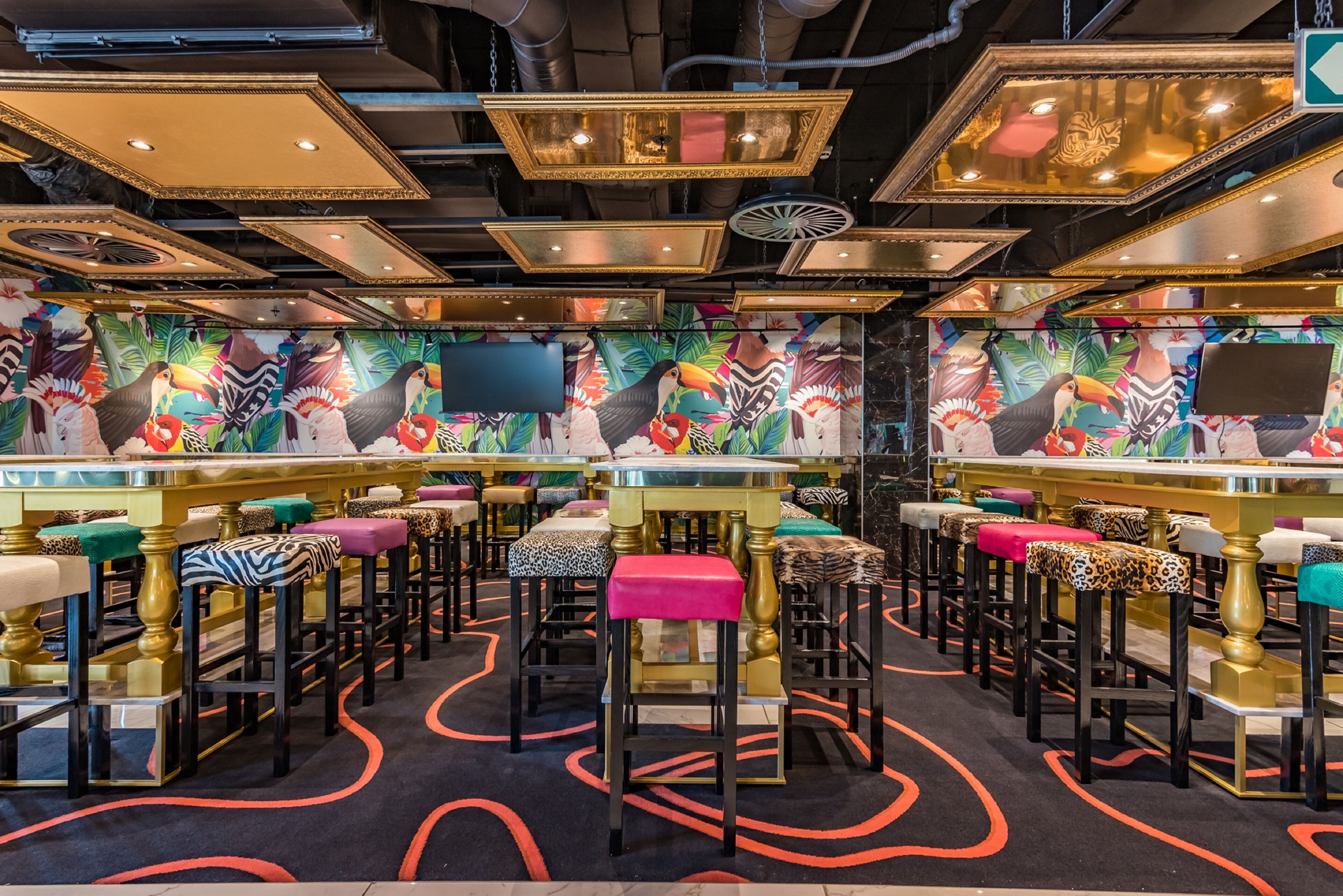 Un área de bar con varios taburetes y mesas, destacada por un mural vibrante con aves tropicales y follaje.