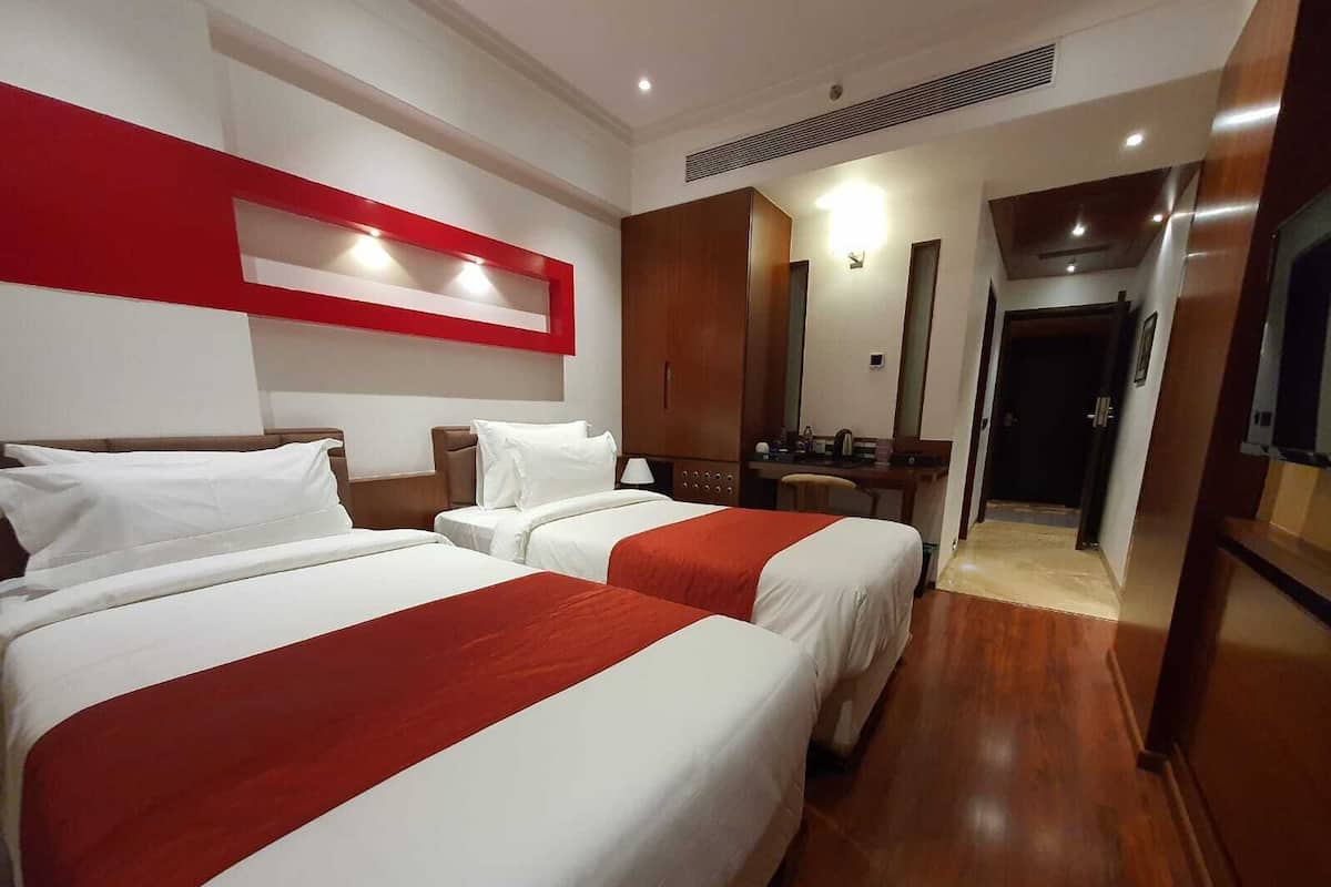 A hotel room with two beds, a wooden floor, a red accent wall, a mirror, and a door leading to a bathroom.